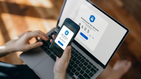 A person holds a smartphone over a laptop to complete a two-factor authentication process. Both screens display security prompts and matching verification codes.