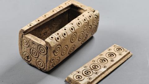 A small box made out of bone with round circles carved into the side for decoration. It is next to a sliding lid that closes the box. 
