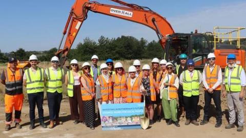 People wearing hard hats and high-visibility vests stand at a housing development site in front of an excavator, holding a sign promoting a council housing project.