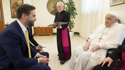 Jd Vance on the left sits facing the pope on the right. Another man wearing a religious outfit stands in the background.