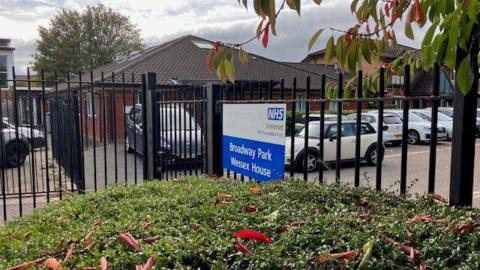 A dark-coloured metal fence behind some bushes, with "Broadway Park Wessex House" written on it in white letters on a blue background. The sign also has the NHS logo on it. Behind the fence and sign are parked cars and also a one-story red brick building.