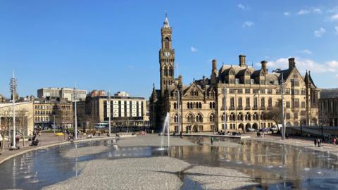 A wide shot of the outside of Bradford City Hall and City Park in front of it with water fountains spraying.