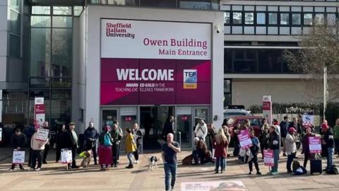 A crowd of people are stood outside Sheffield Hallam University with purple and white strike placards