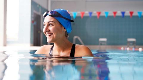 A woman in a blue swimming cap and goggles smiling in an indoor swimming pool.