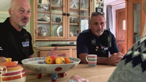 A Trading Standards officer and a police community support officer sit at a kitchen table. There is a fruit bowl and a woman's grey jumper is visible.
