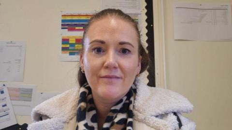 Aisling has brown hair tied up and is smiling at the camera. She has gold earrings on and a leopard print scarf and teddy bear woolly white coat on. 
