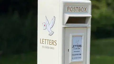 A white post box with the words "Letters to loved ones" on the side below a dove flying with a letter in its mouth. It has the traditional Post Box writing on the front with a little white plaque on the front.