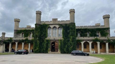 A two-floor gothic style building with green growing up its walls and three large windows at the top and large wooden doors on the ground floor. Two black cars are parked in front of the building next to grass. 