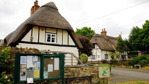 Two thatched houses in Maids Moreton, with half-timbering and cream walls. In the background is a small cluster of trees, while in the foreground can be seen the village noticeboard, with an assortment on posters pinned up.