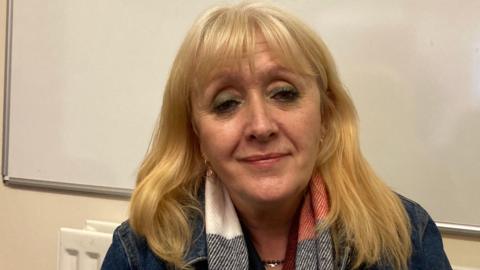 A woman with long blonde hair wearing a red, white and black scarf and a denim jacket, sitting in front of a whiteboard.