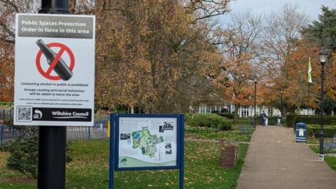 A scene in a Salisbury park showing a Public Spaces Protection Order poster. In the background is an autumnal park scene. 