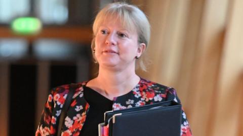 Shona Robison, who has blonde hair, looks to her right while walking towards the camera. She is carrying a black folder held against her chest. She is wearing a red and black floral top. 