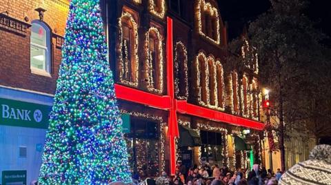 A Christmas tree with lots of blue and green lights. A building nearby has yellow lights and a red bow so it looks like a present. You can see people stood in front of the building.