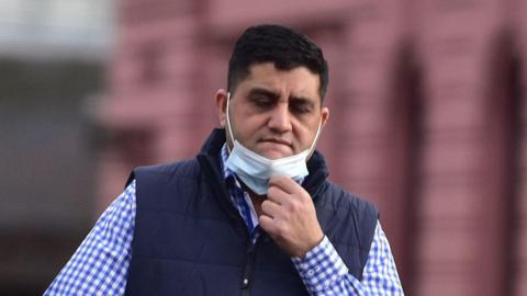 A dark haired man in a check shirt and gilet with a facemask pulled down on his chin