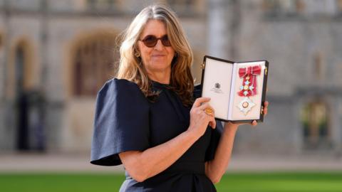 A woman wearing a navy dress with balloon sleeves, stands outside whilst holding an award in her hands.

