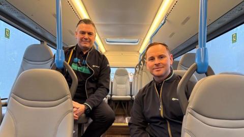 Two men sitting on the bus looking at the camera. They are sitting on different levels of seats and wearing Stagecoach branded clothes. 