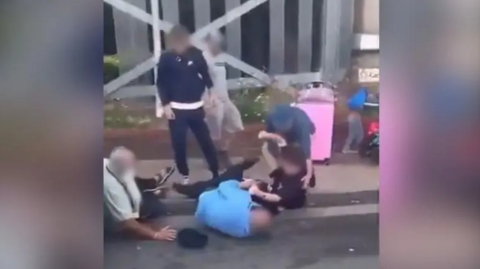 The two men are seen on the floor by a kerb with three other people standing around them and a third man also on the floor. Faces have been blobbed so as not to identify them.