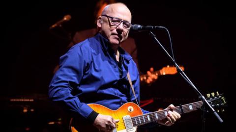 Mark Knopfler performing onstage. He is a bald man with glasses, wearing a blue shirt, and playing a guitar. A microphone is in front of him.