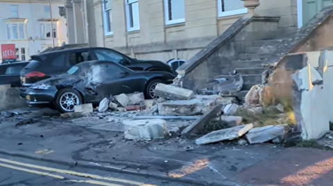A screenshot from a video of the crash taken by a member of the public. It shows several parked cars in a driveway just off the road, with smashed windows and crumpled metal panels. There is also a stone staircase leading up to a green door which has been heavily damaged, with bricks and debris strewn across the pavement.