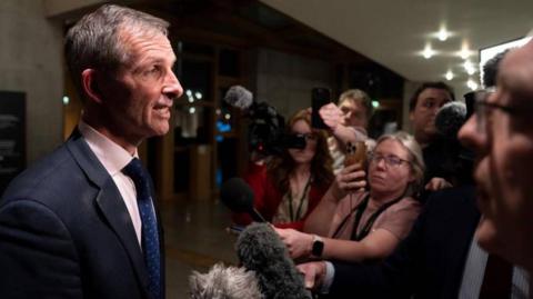 Liam McArthur, wearing a suit and tie, talks to a group of reporters who are holding phones and microphones towards him.
