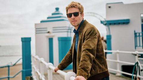 Damien Molony who plays Jim Bergerac stands holding white railings at Lido Jersey, at Havres des Pas. He has short reddish-brown hair ans is wearing dark glasses, a brown suede jacket and blue shirt which echoes the colour of the building behinds him. Gateways to the art deco style lido are painted white with a turquoise edging.