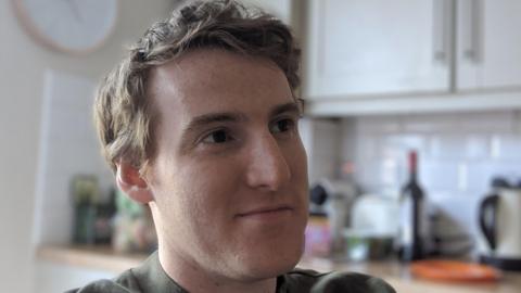 A man is in a kitchen. He has light-coloured, reddish hair and is looking off to the right with a wry, slight smile on his face. He is wearing a green-coloured t-shirt. In the background, white kitchen cupboards and appliances can be seen