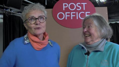 Jo wears a blue jumper and pink shawl. She wears black rimmed round glasses and has short grey hair. Pam wears a turquoise fleece with a grey polo neck underneath. She has grey hair in a bob and is smiling at Jo as she speaks.