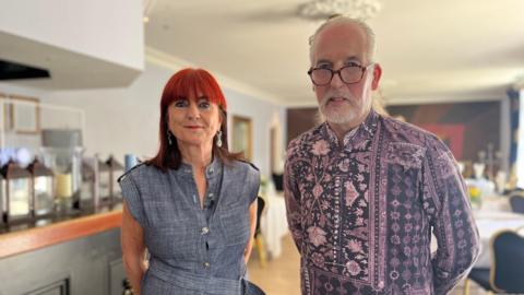 Steve Cuddy and Gillian Etherdige stand together in the dining room of their hotel. To their left is a bar and behind them are tables and chairs with white tablecloths on top. Etheridge has bright red hair and wears a denim dress. Cuddy has grey hair and beard, is wearing glasses and a purple and navy shirt.