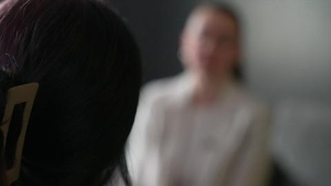 The back of a woman's head. She is speaking to a woman with dark hair and a white suit jacket 