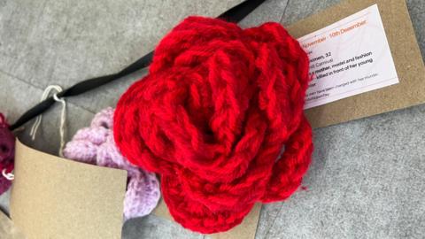 A crocheted red rose. Underneath it is a card with information about a woman (most of it not visible).