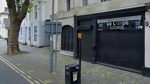 A night club with black shutters covering the entrance and front of the premises. There is a bin and parking sign on the pavement, as well as a lamppost and further signage. There is a tree and further buildings visible in the left hand side of the image.