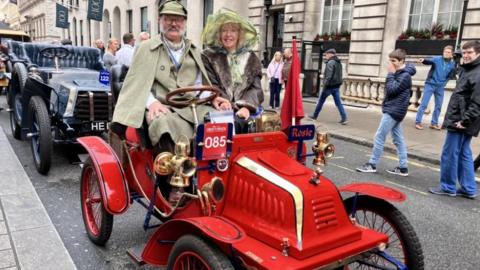 A man and woman, dressed in fashions from the early 20th Century, sit atop a red car from the same period.