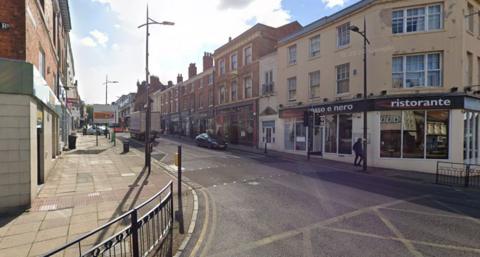 A city-centre street, lined by shops and restaurants, with lorries and cars driving down it and signage by the side.
