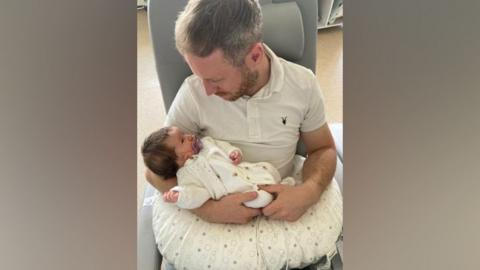 Pete Grimwade pictured sitting down holding his baby daughter, Isabella, in his arms, looking down at her while she looks up at him