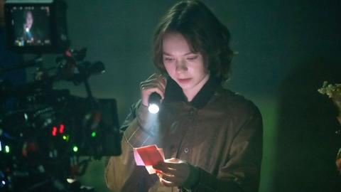 A young woman with a brown bob, wearing a dark green wax jacket. She in in the dark and her face it lit up by the torch she is holding. She is reading small squares of paper. There is a film camera in the foreground to the left.