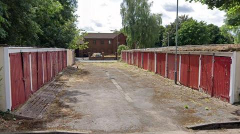 Two rows of garages on a housing estate. They have faded red doors and slightly sloping roofs. 