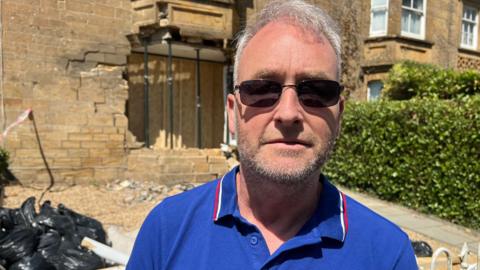 Mark Talbot-Phillips is standing outside his house which has a big hole in it. He is wearing a blue shirt and sunglasses.