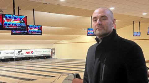 Jonathan Ruff is wearing a dark coat and jeans stands inside a bowling alley near a railing that overlooks several empty bowling lanes. Multiple overhead screens display colorful graphics above the lanes, and seating areas with bowling ball returns are visible throughout the space. The lighting is warm, and the setting appears clean and modern.