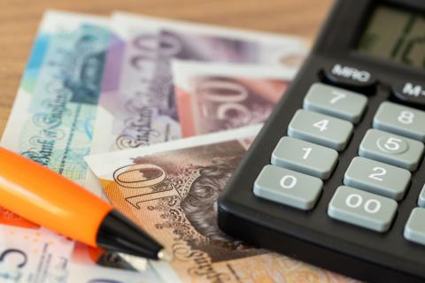 Bank notes of varying amounts are laid overlapping on a table, and on top, is an orange pen and a large calculator.