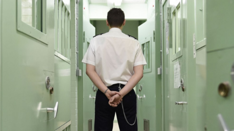 The back of a prison warden is shown, standing in a corridor of mint green prison doors