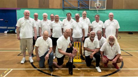 Players from Bexhill Walking Cricket, with a team photo around the wickets inside a sports hall, wearing white tops. 