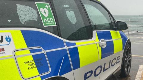 A Jersey Police car is seen parked up with the sea visible beyond car park railings 
