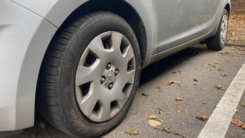 Two dirty looking black tyres with grey rim caps on a silver car parked in a car park