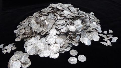 A huge pile of silver pennies on a black background. Some of the coins have spilled into smaller clusters around the main pile and some look bent out of shape or are broken in half.