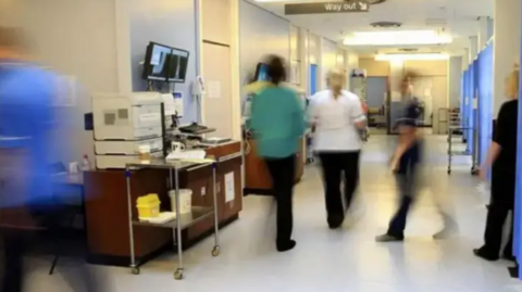 A generic image of medical staff, who have been blurred visually, moving around a hospital attending to patients. They are dressed in various types of medical scrubs and can be seen walking through a ward which has patient areas on the right-hand side.