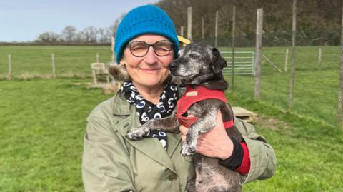 Mole is being held by her owner Zara who is standing on a green field of grass. She is a small dog with fur that is a mixture of black and grey