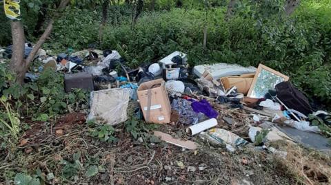 A pile of fly-tipped rubbish spread across the edge of a verge. There are boxes, rolls of foam, a mattress, building waste and black plastic bags of waste. Behind it are trees and nettles. 