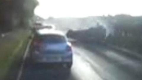 A grainy still from Dascham showing a country road, with a line of traffic on the left and a car skidding on its roof along the right hand side.