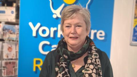 A woman with short grey hair stands in front of a banner with the Yorkshire Cancer Research logo on it. She wears a dark green top and a green and pink leopard print scarf.