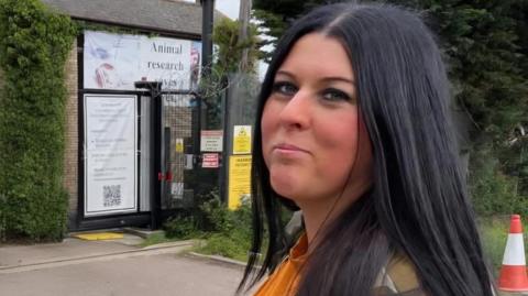 A woman with long black hair standing outside near a green hedge, looks sideways towards the camera